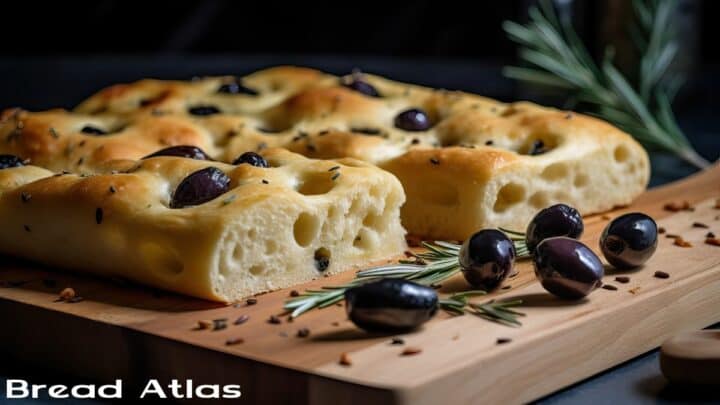 Focaccia on a wooden board with olives and rosemary in the background.